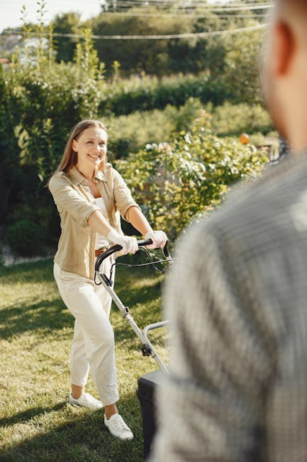 A woman is engaged in garden maintenance, pushing a lawn mower across a well-kept grassy lawn in a backyard. She has a cheerful expression, dressed in a beige jacket and light-colored trousers, with s