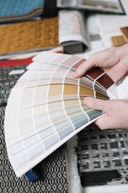 A person holding and flipping through a large garden color palette sample book featuring various shades of earth tones, beiges, browns, and greens, in a workspace with fabric samples and design materi